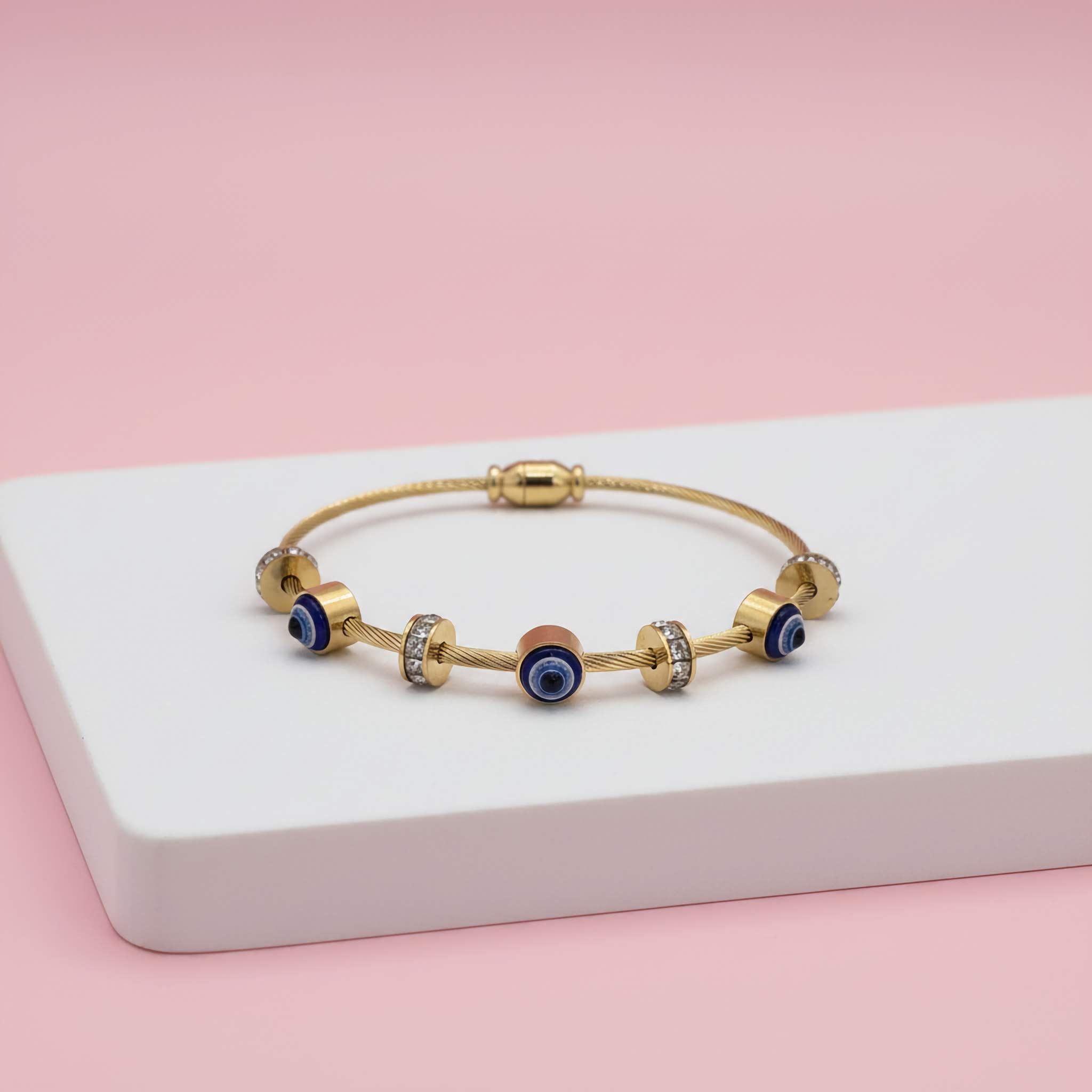 18K gold plated bracelet with blue evil eye beads on a white platform with a pink background