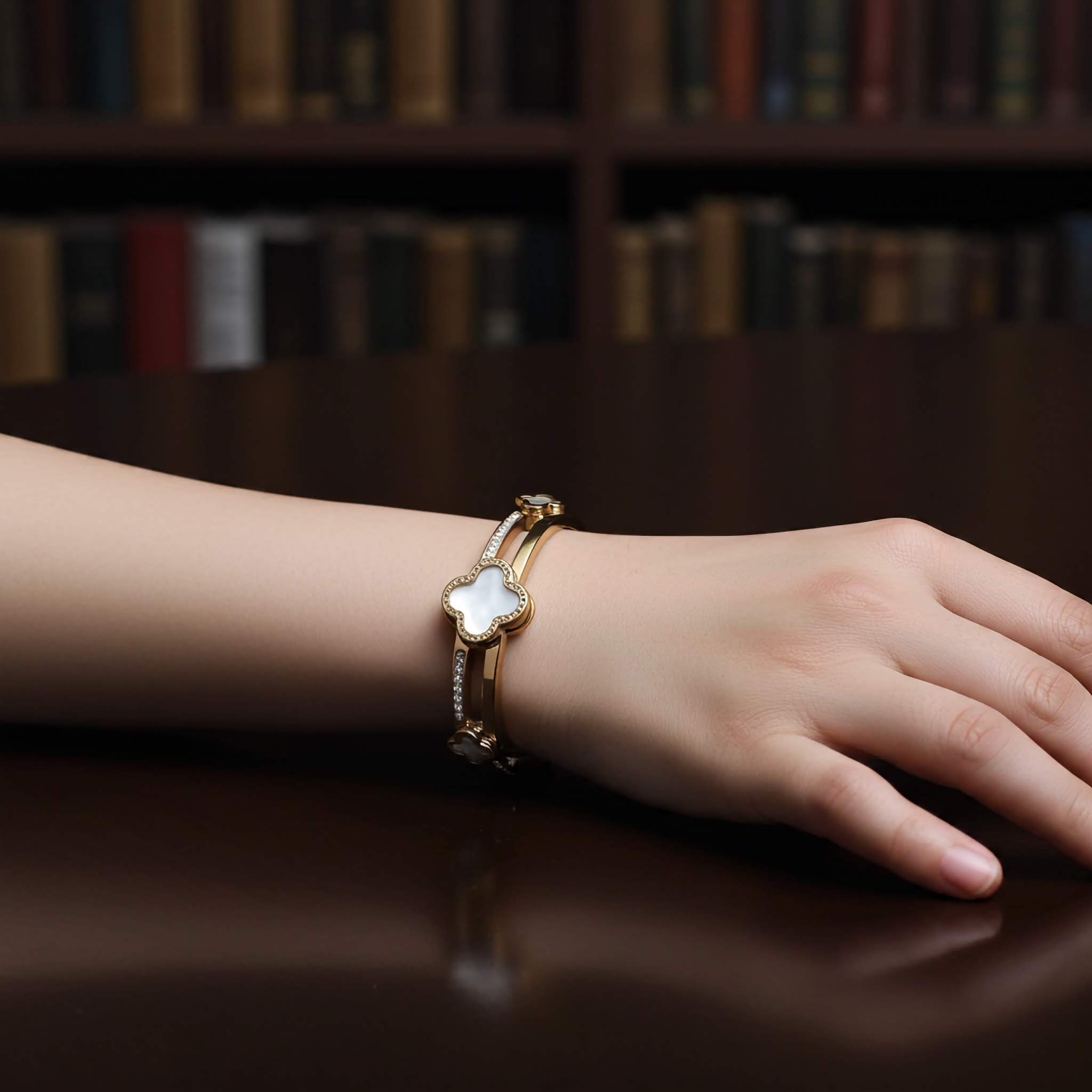 Hand wearing a gold clover bracelet with a white flower charm on a wooden surface with bookshelf background