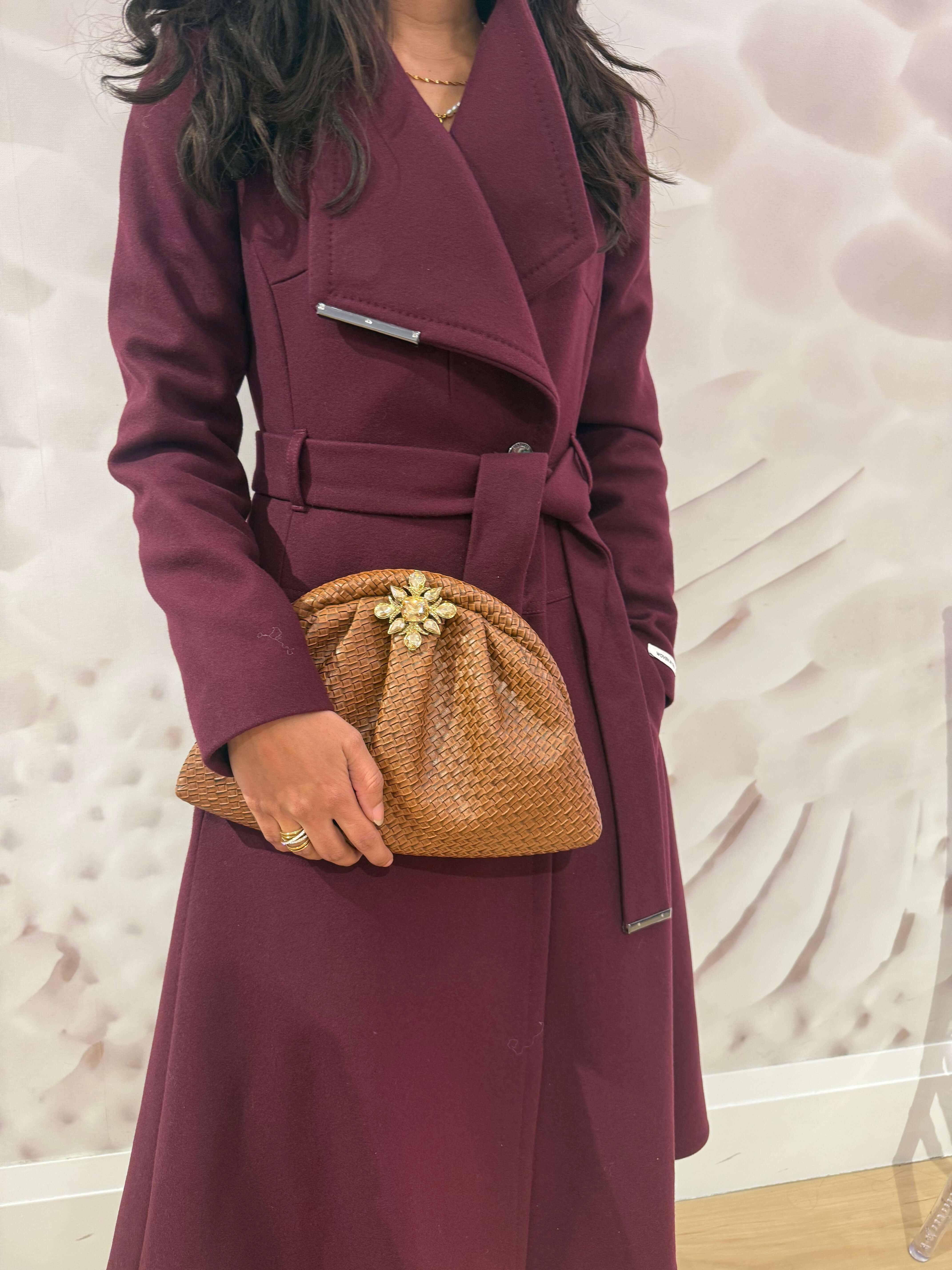 The Luxe Weave Clutch held by a woman in a burgundy coat, featuring a textured caramel vegan leather design and floral embellishment.