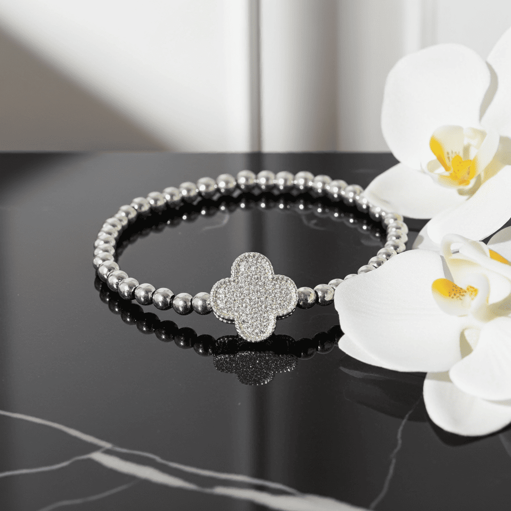 Silver beaded slip on bracelet with clover-shaped clasp on a black surface with white flowers.