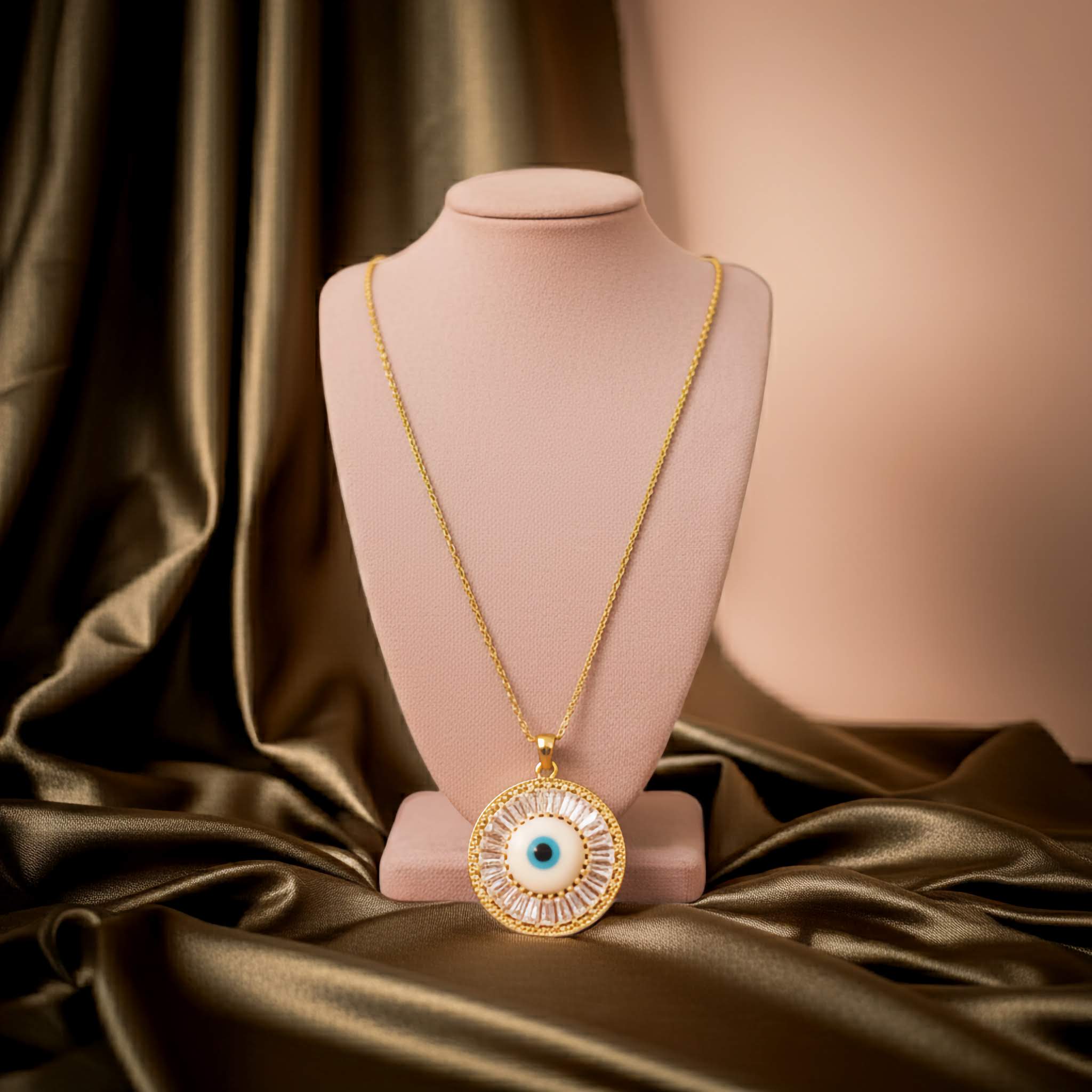 Gold necklace with an eye pendant on a pink mannequin bust against a brown fabric background