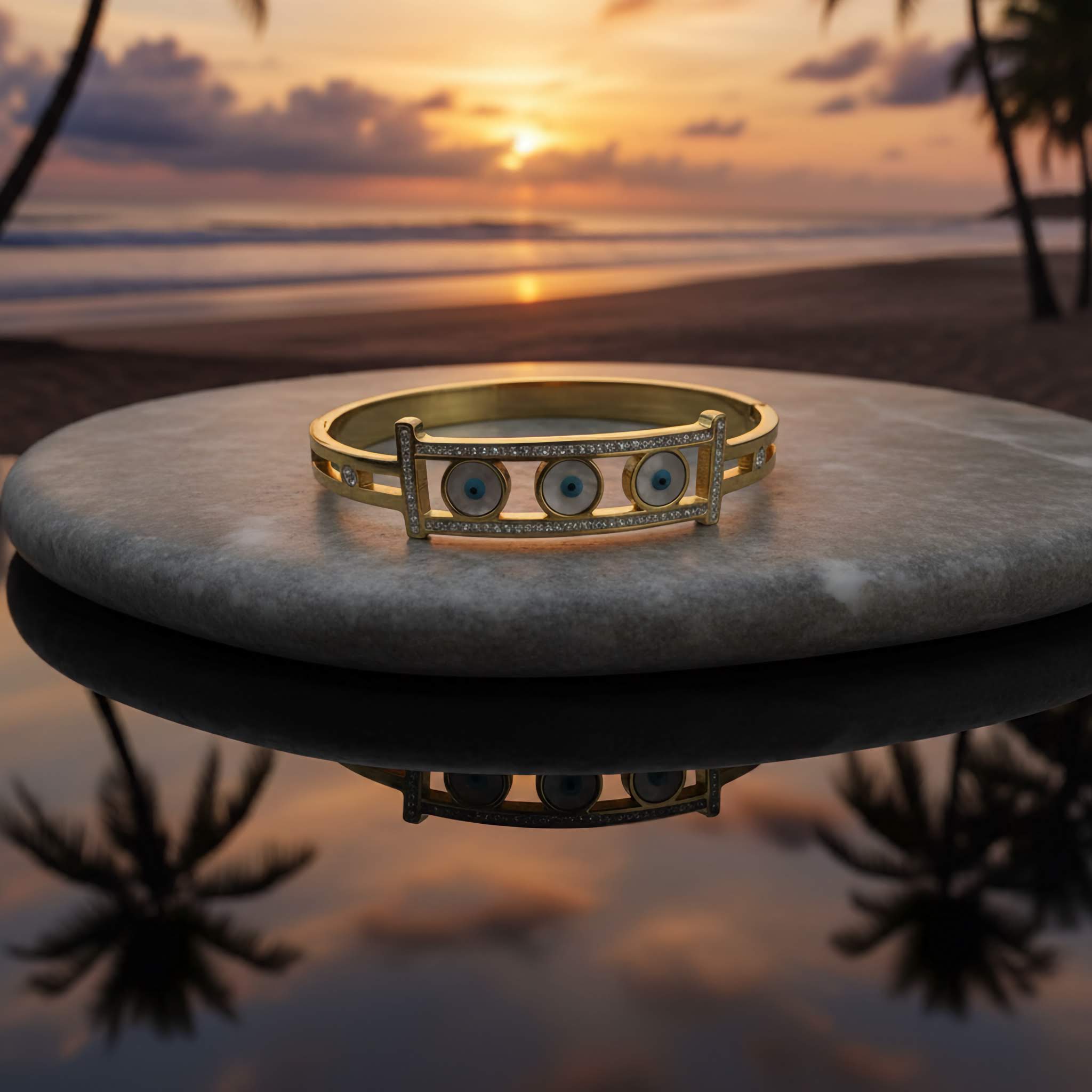Gold bangle bracelet with blue evil eye stones on a reflective surface at sunset