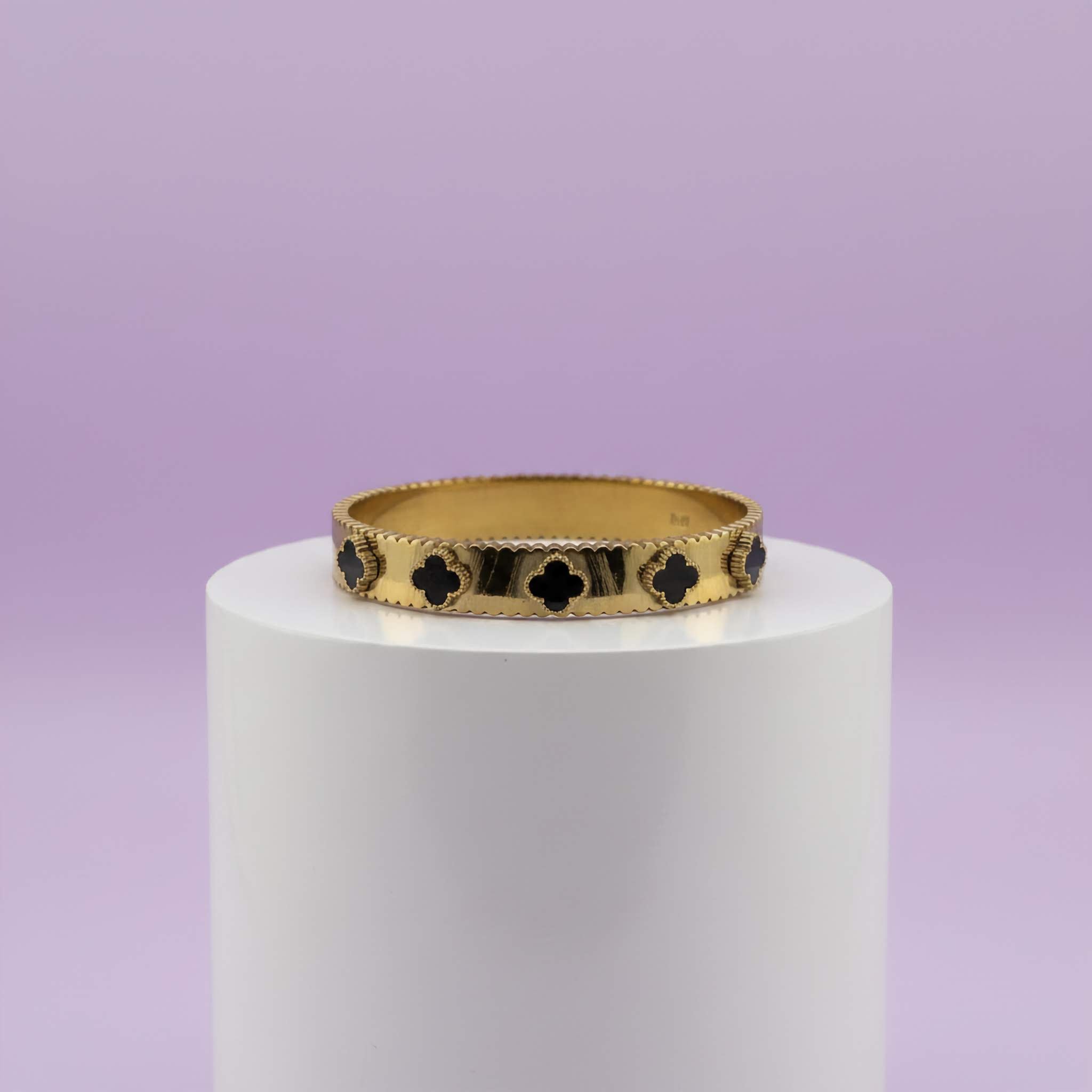 Gold bracelet with black clover patterns on a white cylindrical stand against a purple background