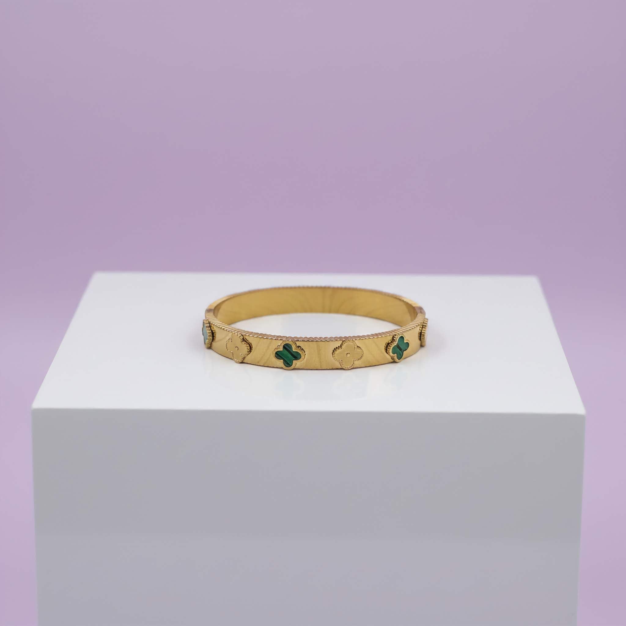Gold bracelet with green clover leaf on a white cube against a purple background