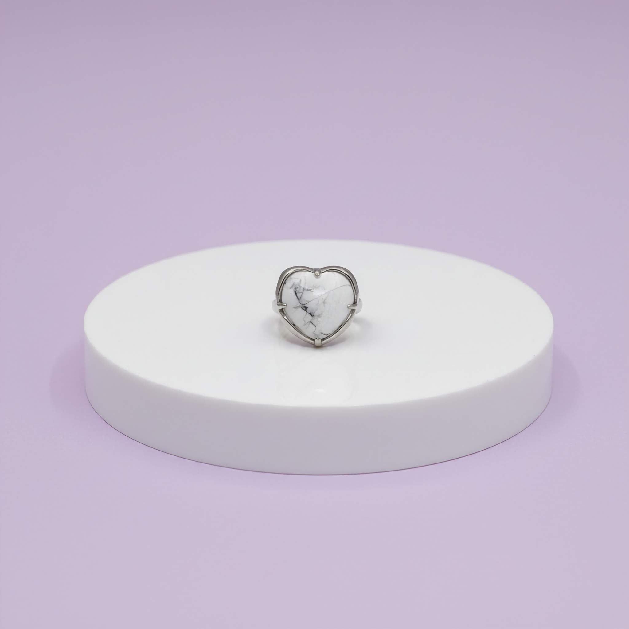 Heart-shaped silver ring on a white stand against a light purple background