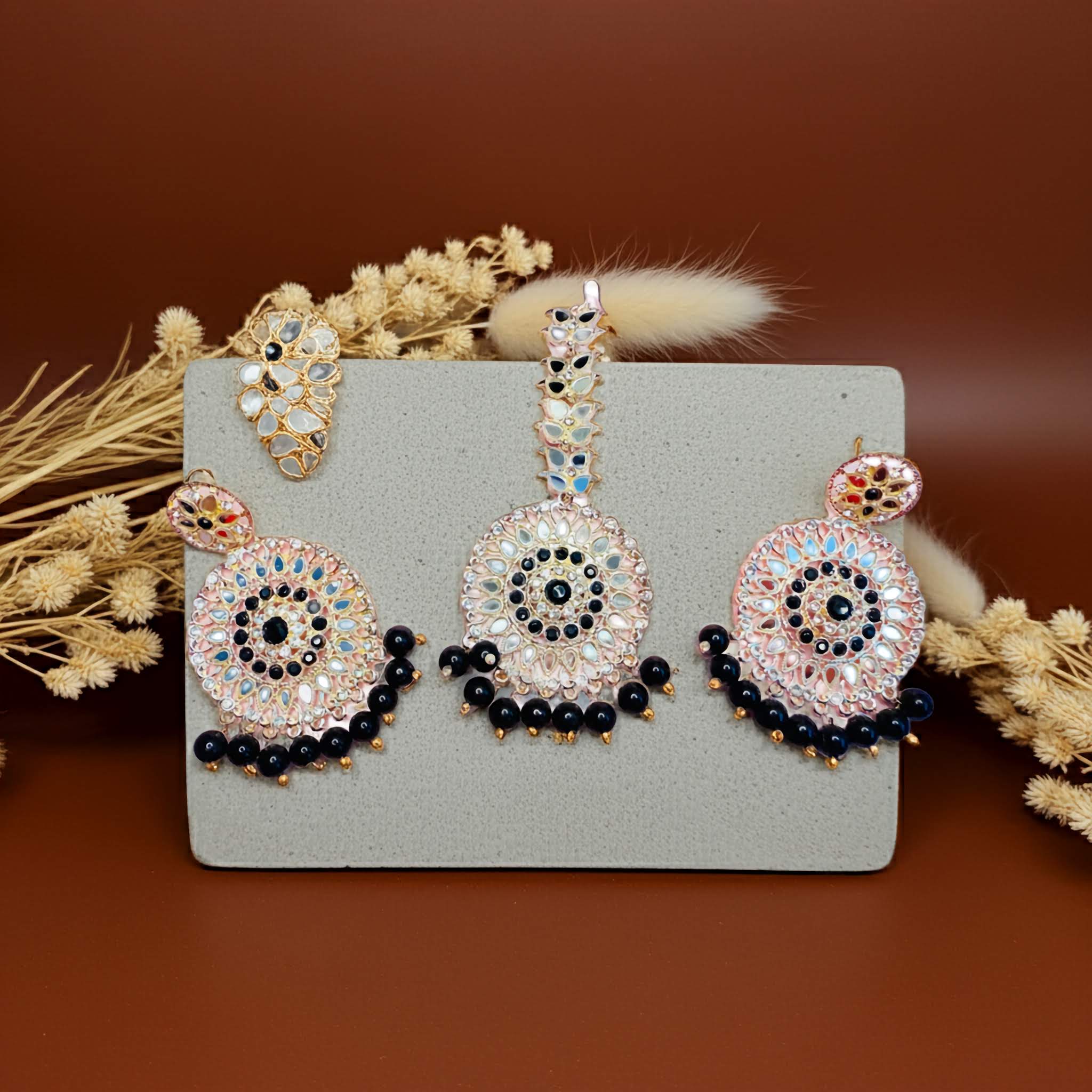 Mirror Decorative earrings and tikka with black pearls on a gray card with dried flowers on a brown background