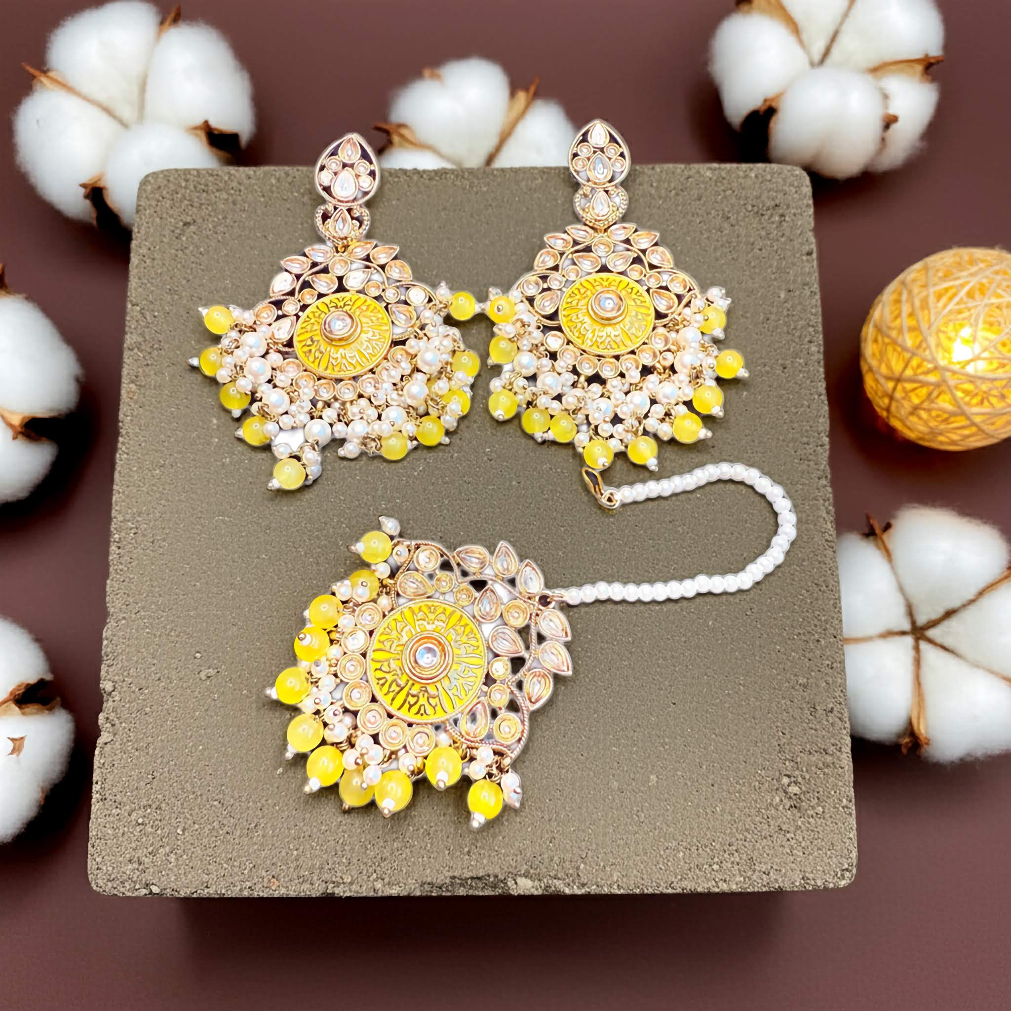 Mirror design earrings and tikka set with yellow and gold elements on a textured surface with cotton flowers in the background.