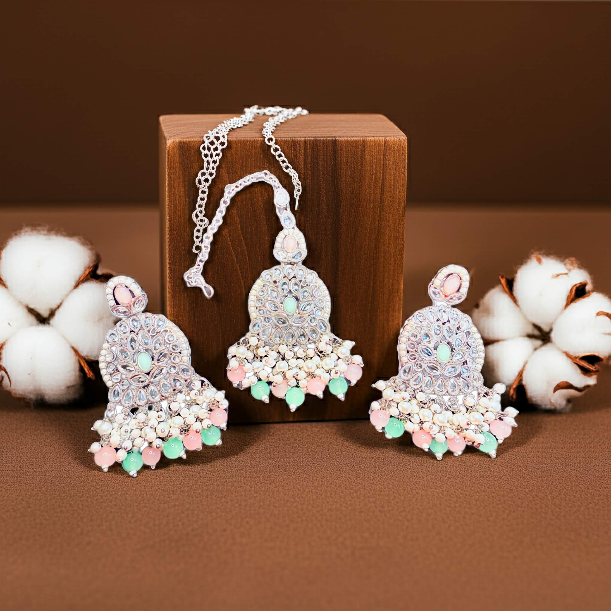 Mirror designed Decorative earrings adn tikka with pink and green accents on a brown surface with cotton balls.