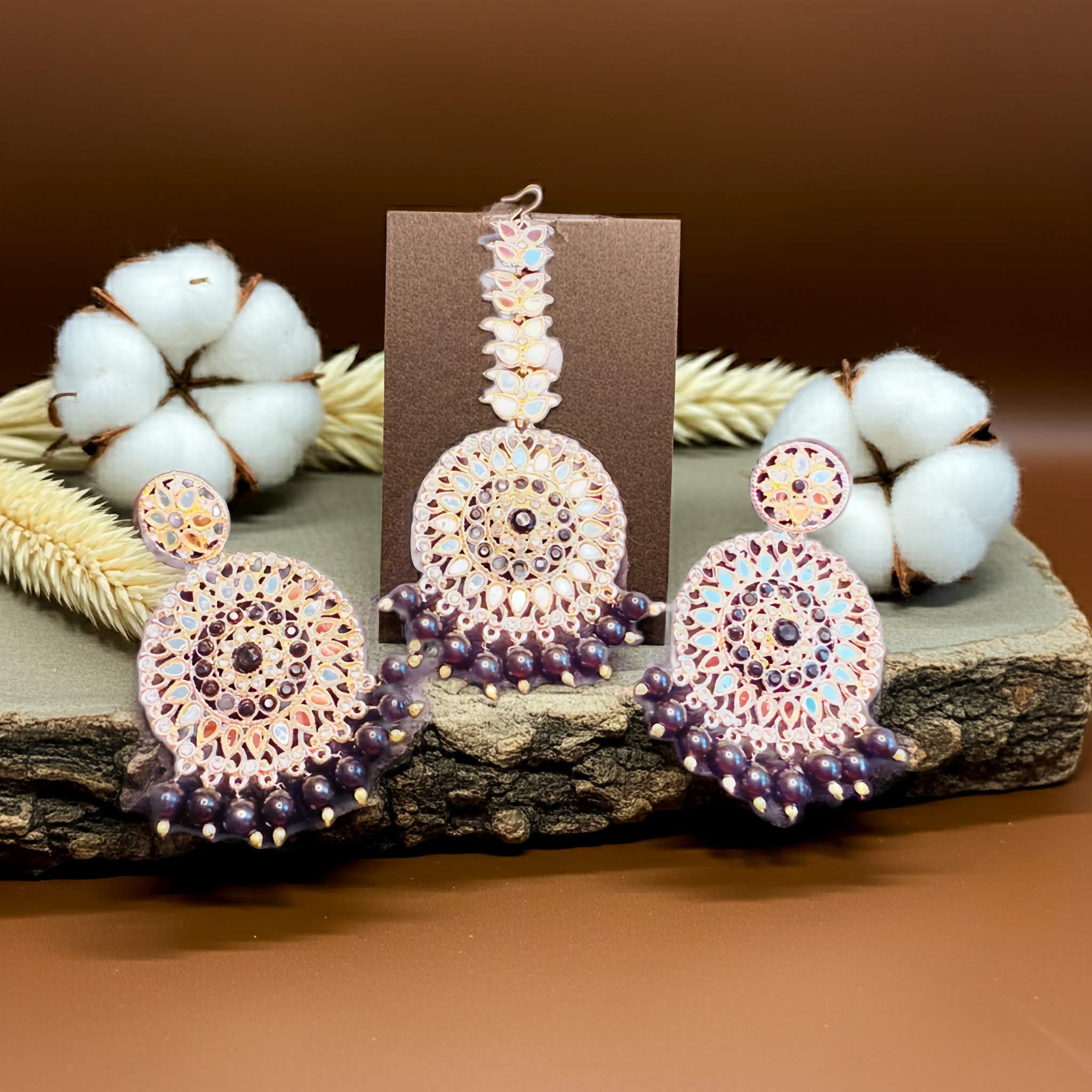 Ornate mirror work earrings and tikka with purple and pink gemstones on a wooden surface with cotton and straw.