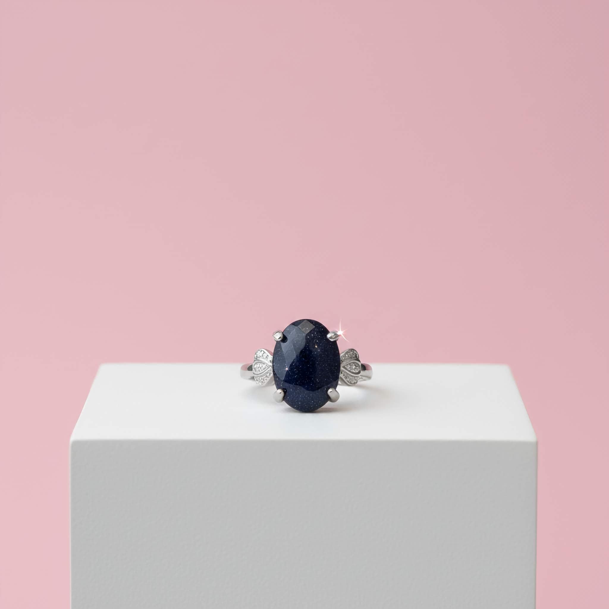 Ring with a large dark sparkly blue gemstone on a white pedestal against a pink background
