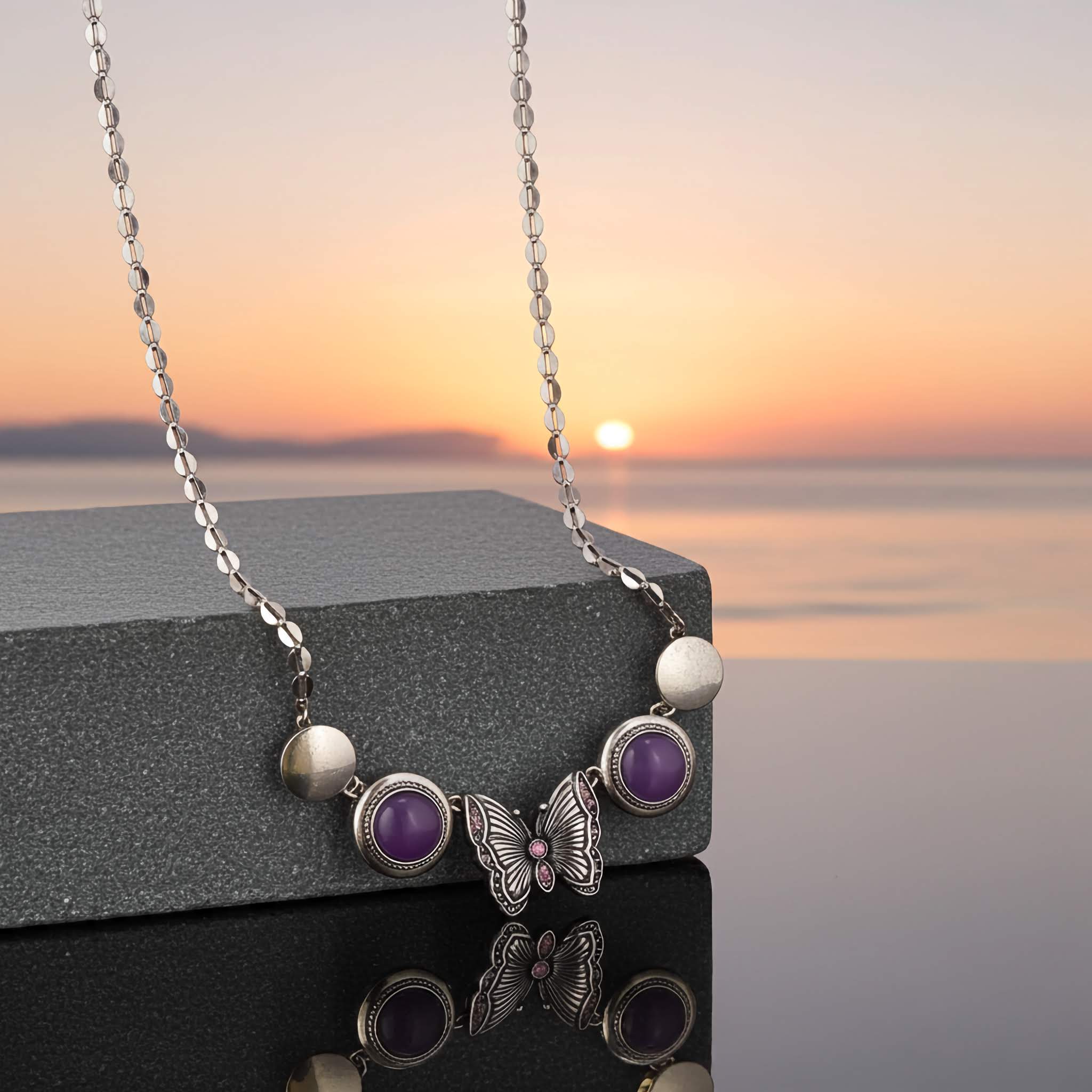 A Silver tone Necklace with butterfly and purple bead pendant on a reflective surface with sunset in the background