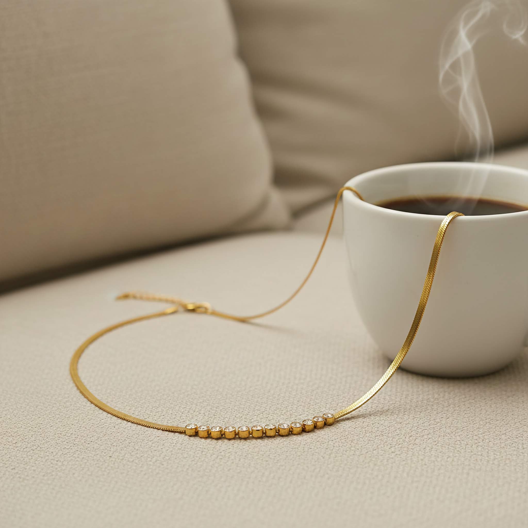 Gold plarted beaded necklace on a beige surface with a steaming cup of coffee in the background