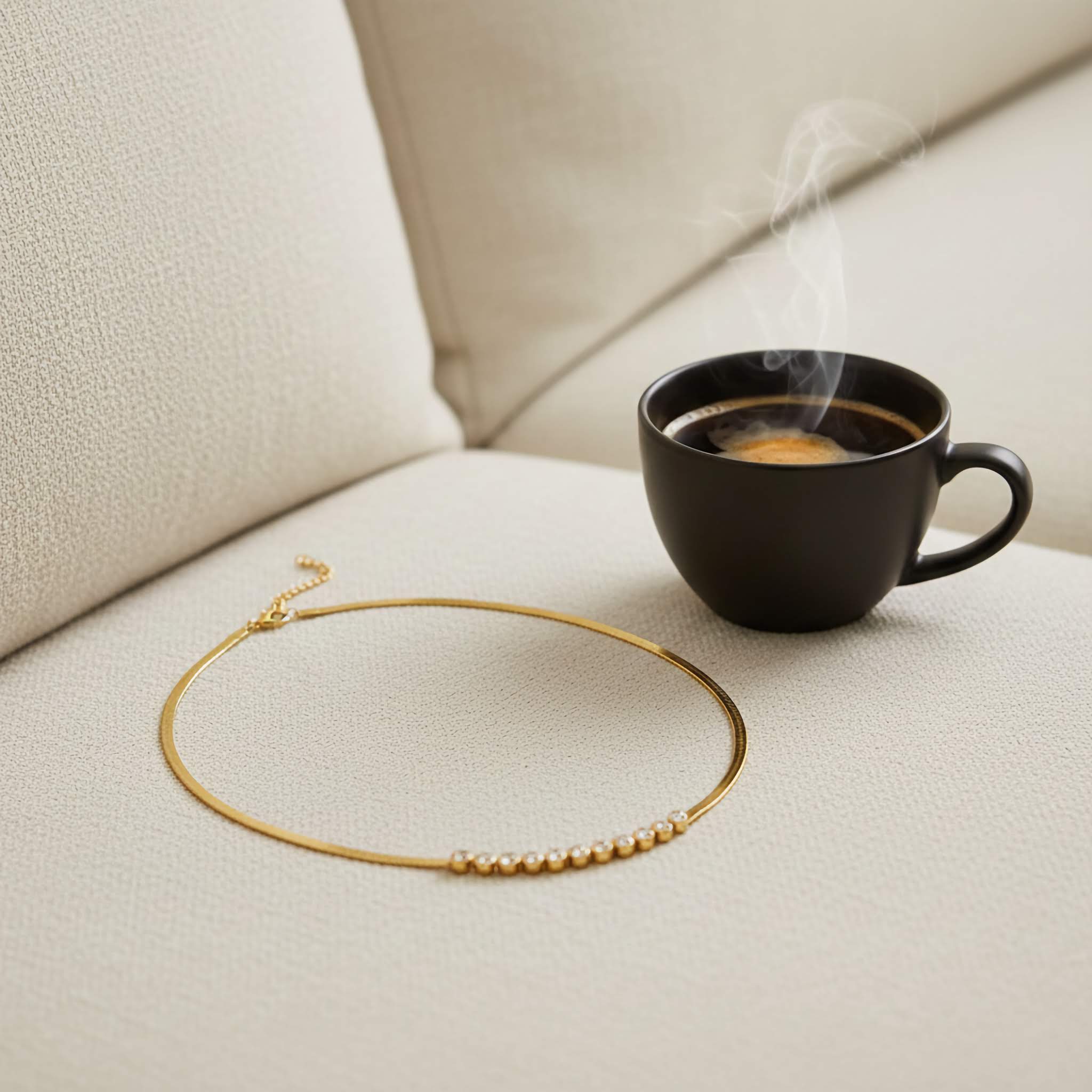 Black coffee cup with steam on a beige surface next to a gold necklace of clear zirconia diamonds.