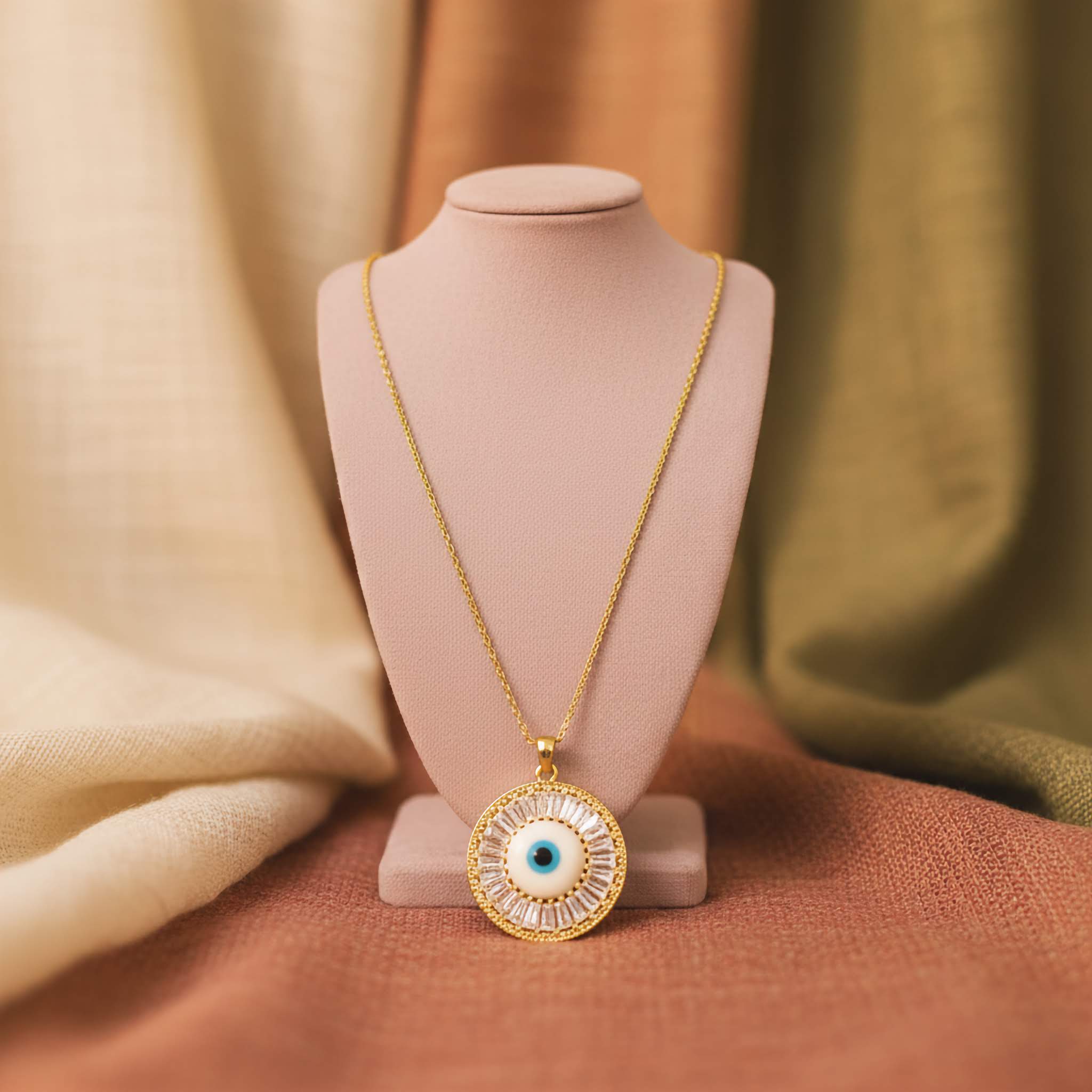 Gold necklace with an eye-shaped pendant on a pink stand against a soft, multicolored fabric background