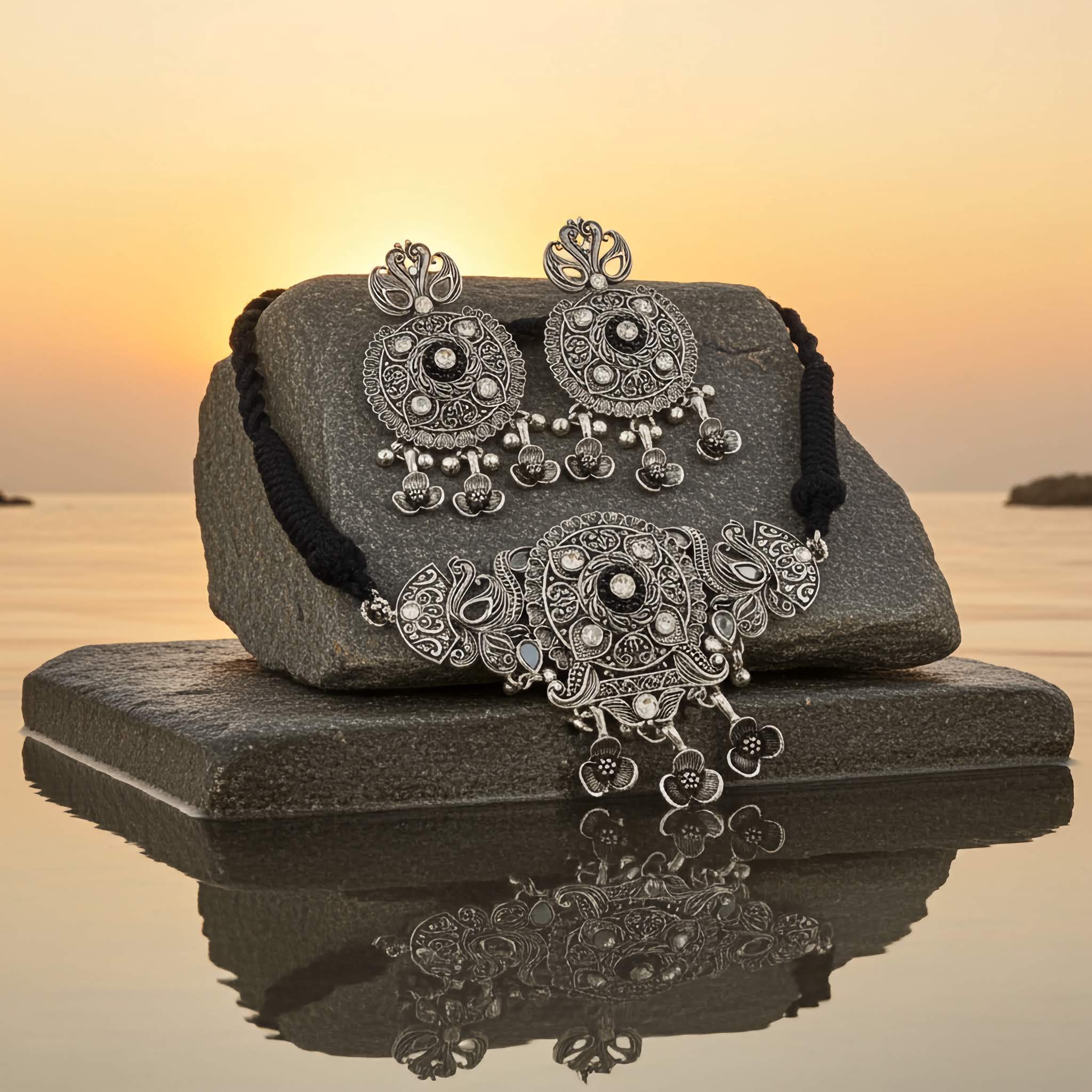 Ornate silver choker set with jhumka earrings on a reflective surface with a sunset background