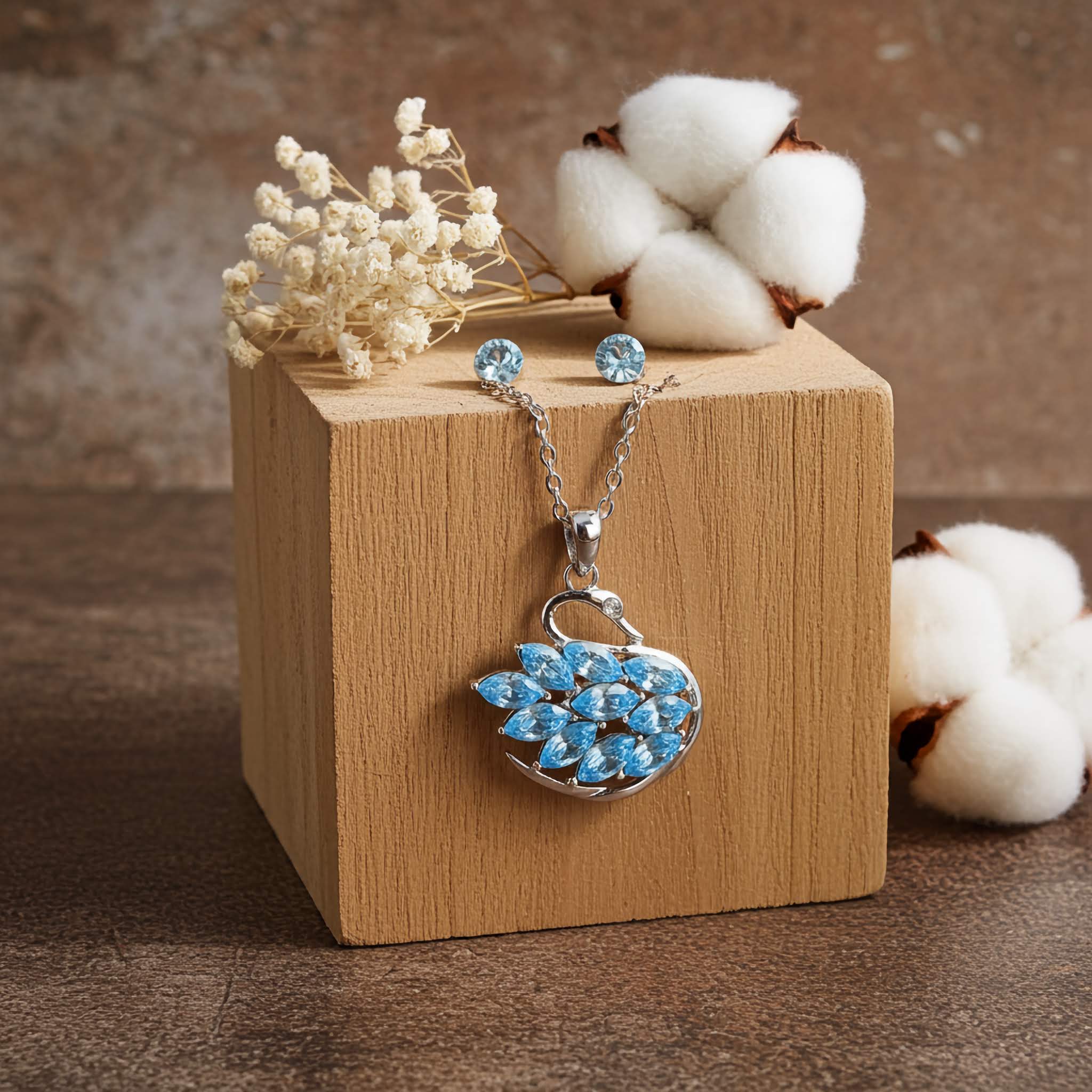 A silver Jewelry set with blue swan pendant and earrings on a wooden block with cotton and flowers.