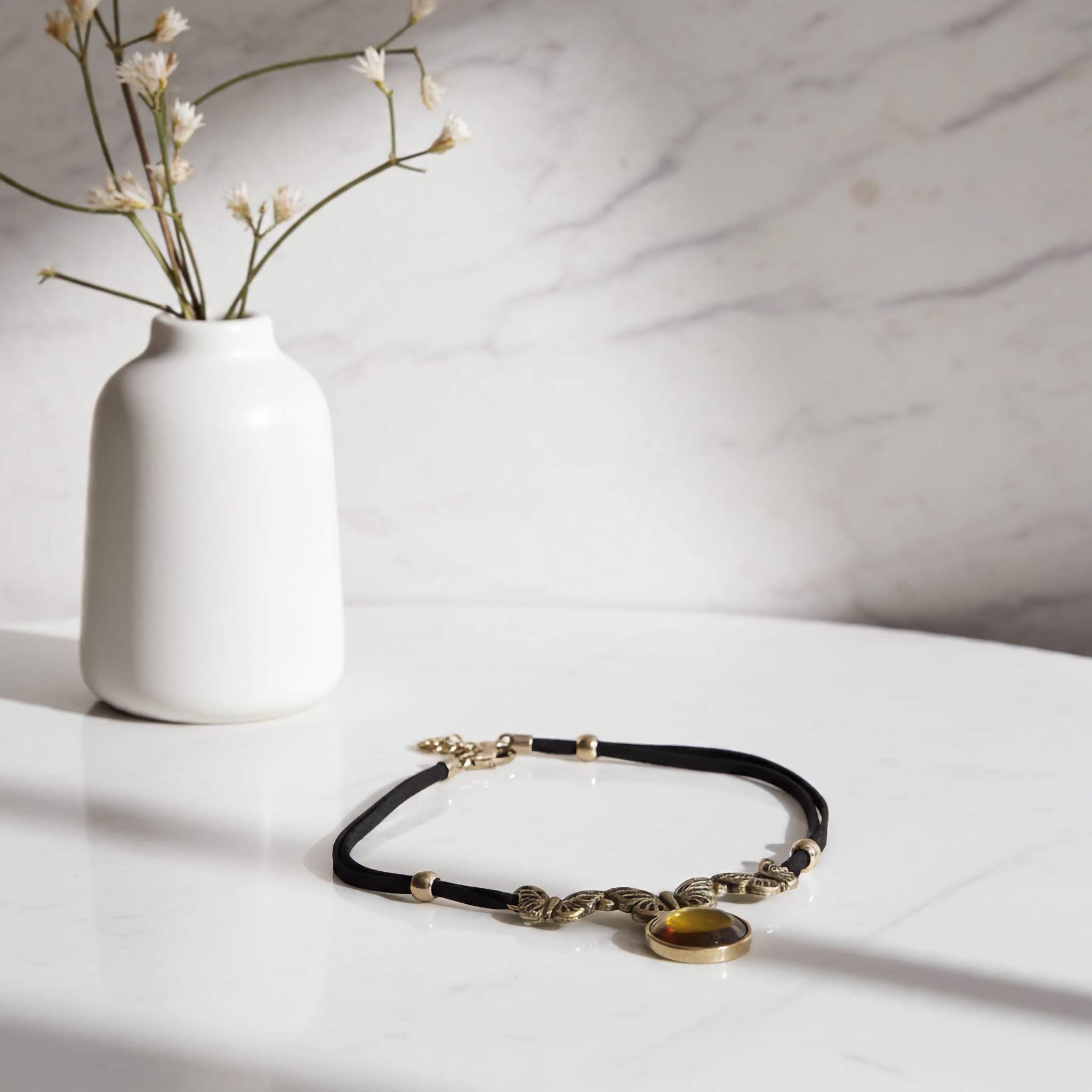 Black choker necklace with gold accents on a white surface, next to a white vase with dried flowers.
