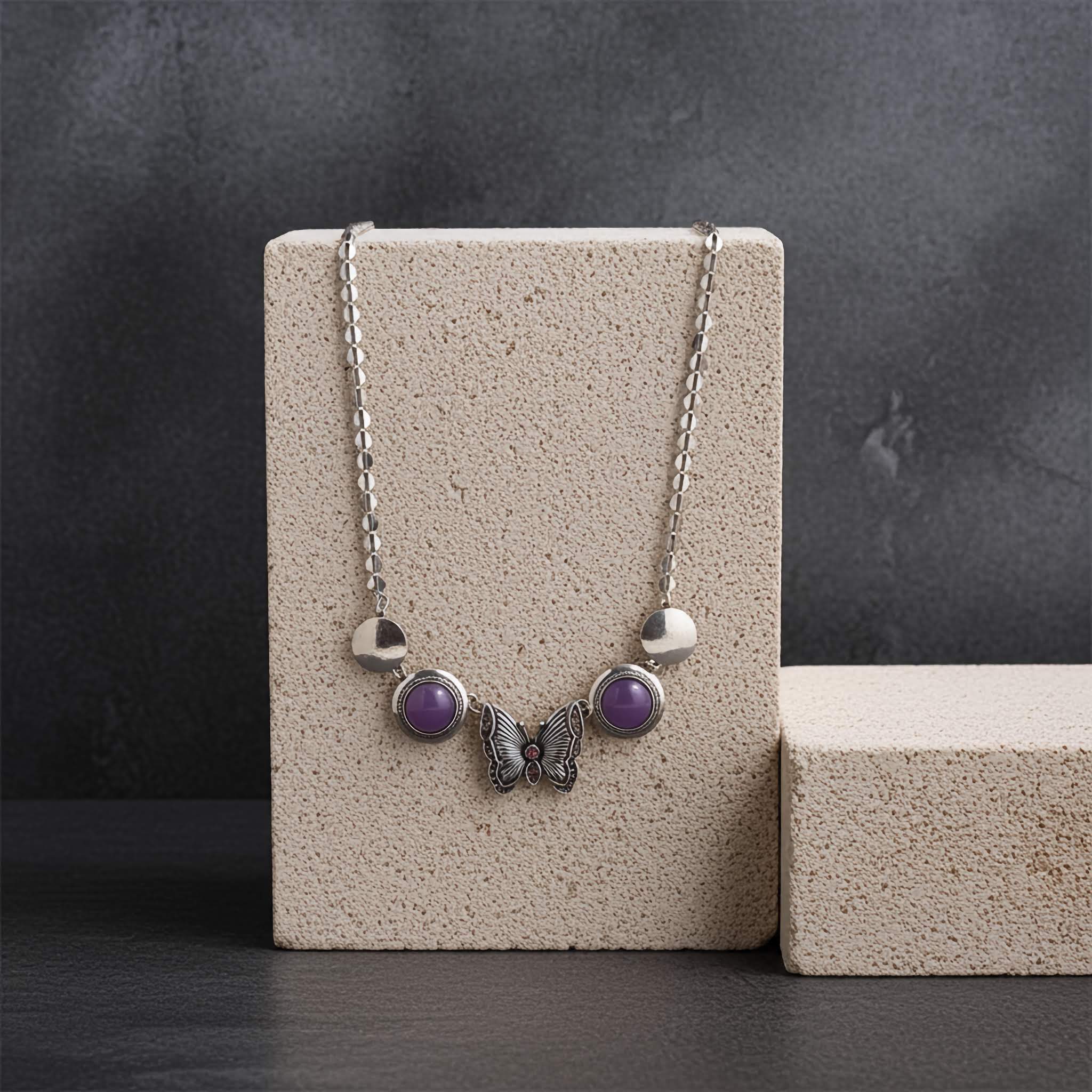 A silver tone Necklace under $10 with purple stones and a butterfly pendant on a beige stand against a dark background