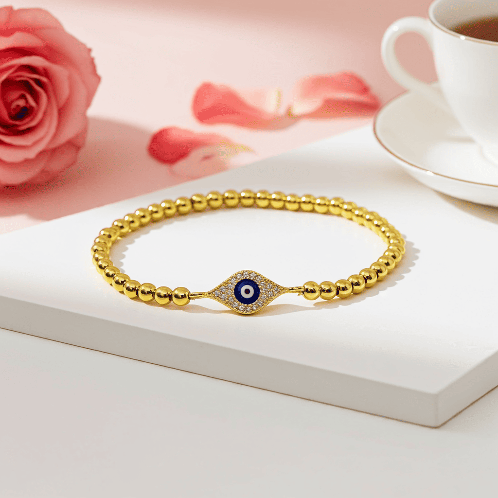 Shop Evil eye slip-on Gold bracelet with a blue evil eye charm on a white surface with a pink rose and tea cup in the background.