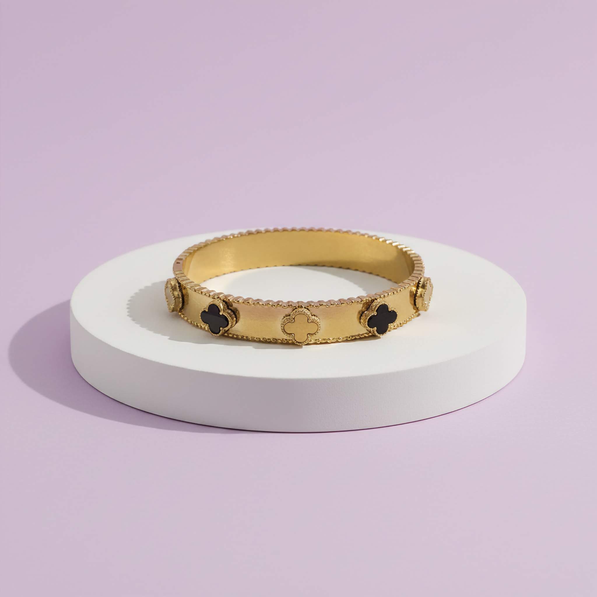 Gold bracelet with black clover designs on a white stand against a light purple background