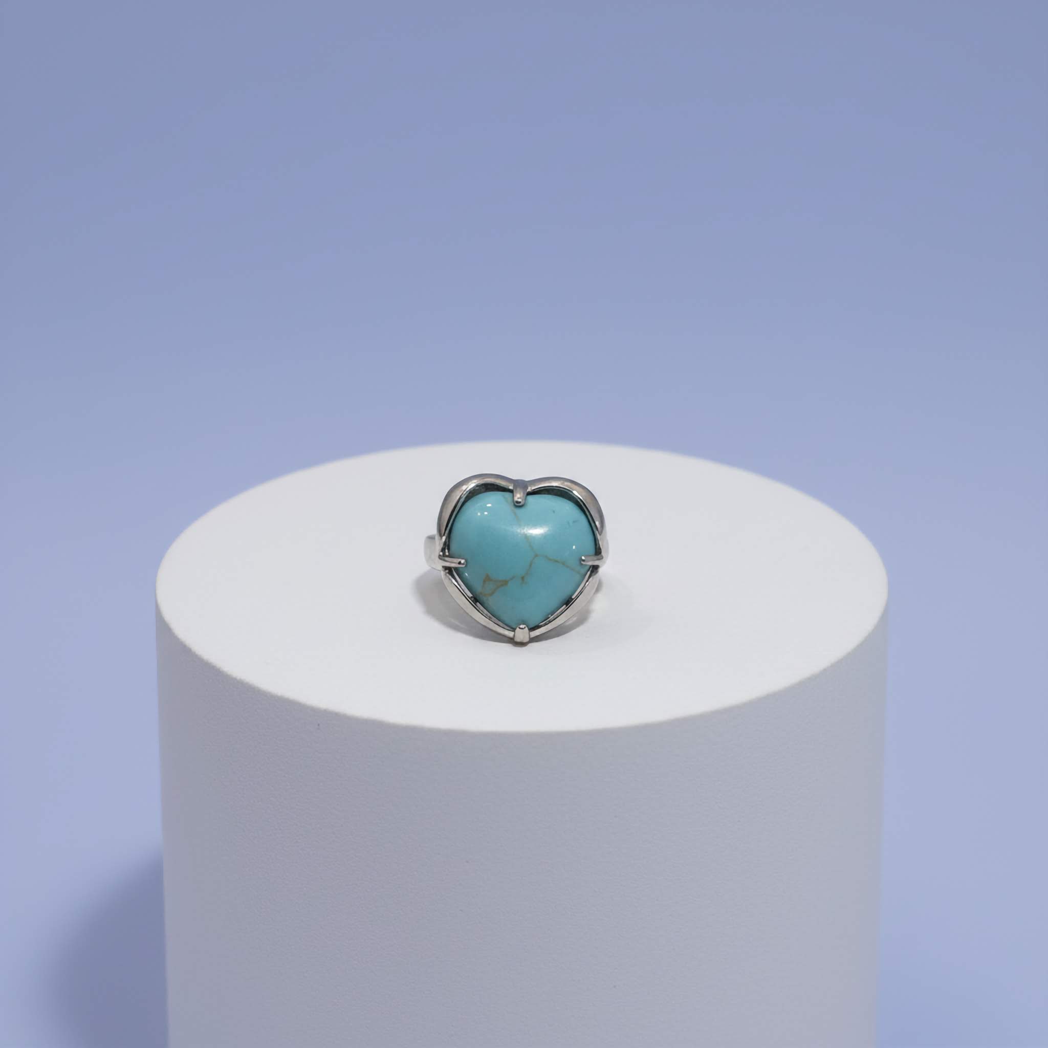 Heart-shaped turquoise stone ring on a white cylindrical stand with a light purple background
