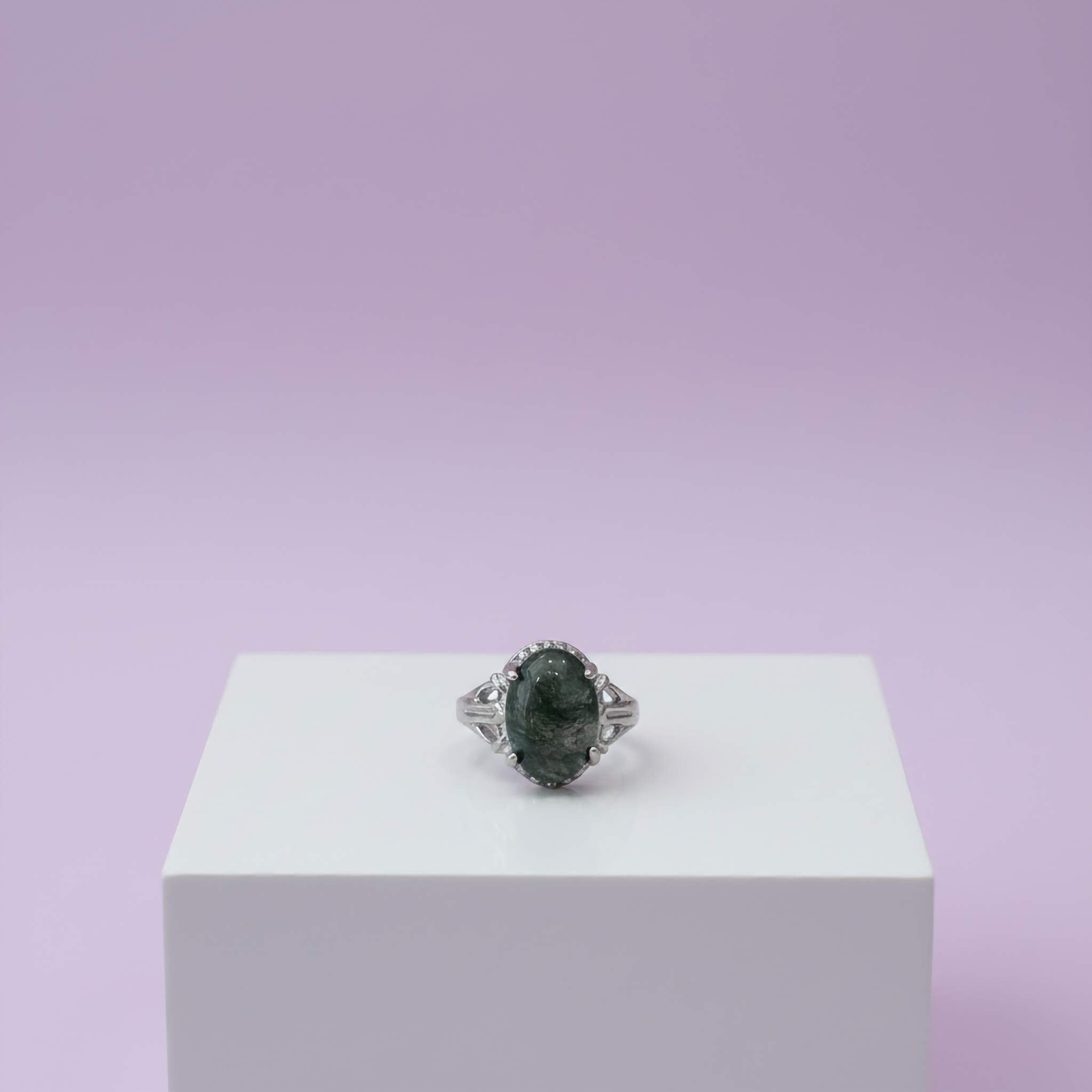 Silver ring with a dark green gemstone on a white pedestal against a light purple background