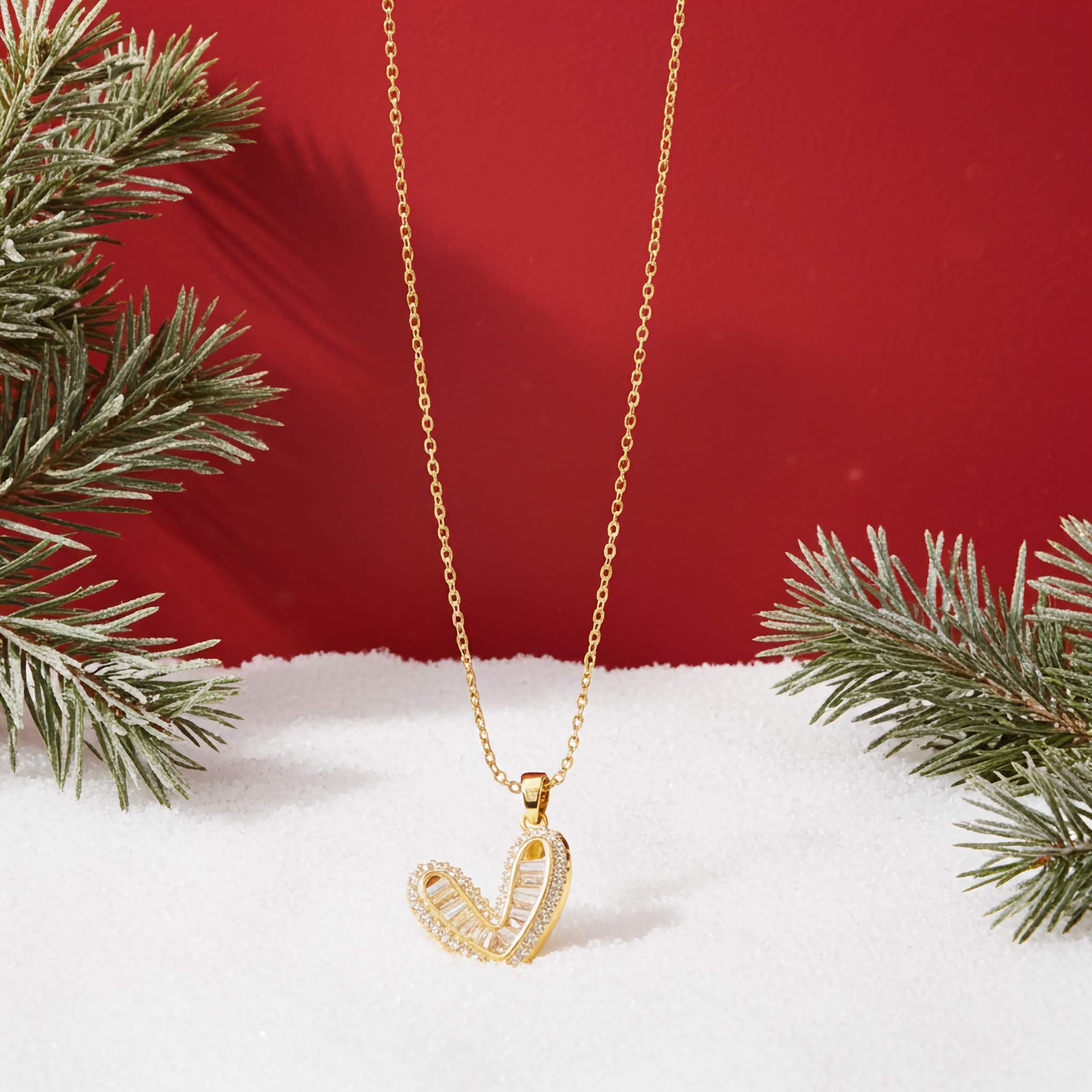 A Gold necklace with a heart-shaped Swaroski pendant on a red background with snow and greenery.