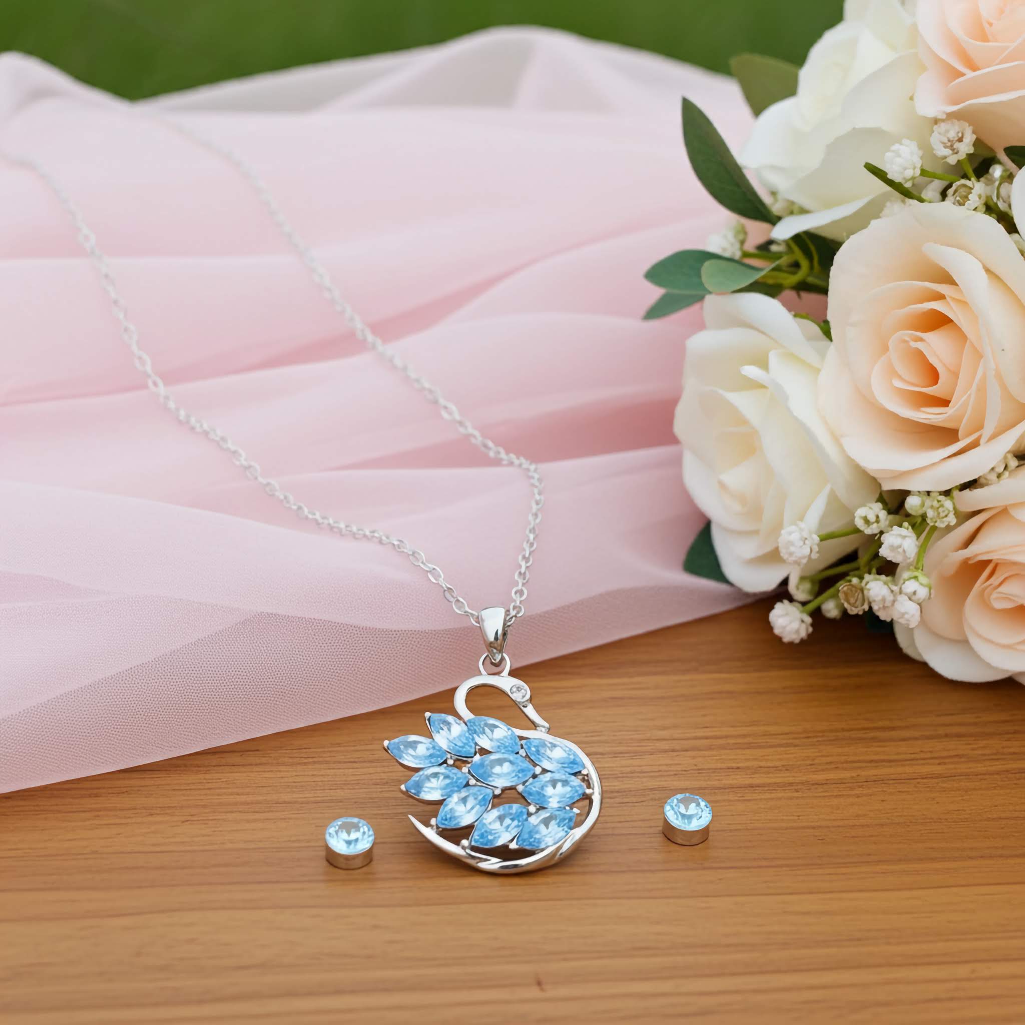 A Swarovski silver swan Jewelry set with blue gemstone pendant and earrings on a wooden surface with pink fabric and flowers.